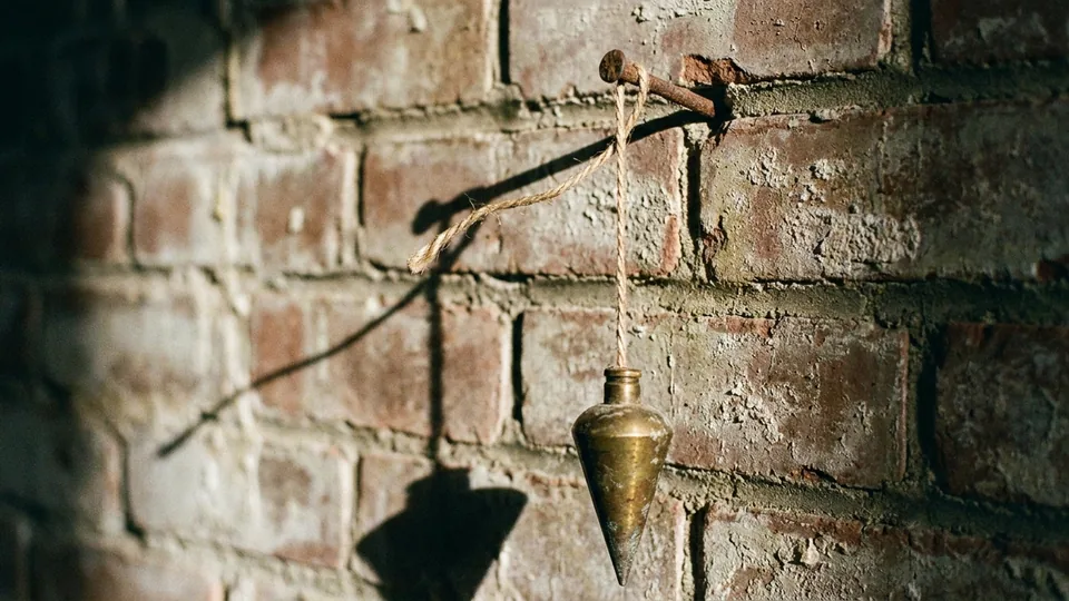 A brass plumb bob hangs from twine tied to a rusty nail hammered into a weathered brick wall, casting long shadows in sunlight.