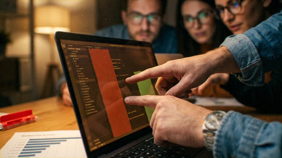 Several adult colleagues (at least three visible, two wearing glasses) clustered around one laptop; one person in a rolled denim shirt with a metal wristwatch points at the screen while another finger also indicates the display; the laptop runs a code editor.. Coworkers.